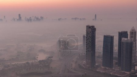Téléchargez les photos : Grande jonction de carrefour entre le quartier JLT et la marina de Dubaï recoupée par Sheikh Zayed Road nuit à jour transition aérienne timelapse. Trafic automobile couvert par le brouillard matinal - en image libre de droit