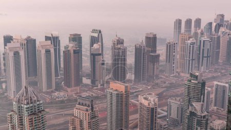 Téléchargez les photos : Les gratte-ciels et les tours de marina de JLT près de Sheikh Zayed Road se succèdent de nuit au jour. Bâtiments résidentiels éclairés et skyline avec villas. Foggy matin avant le lever du soleil - en image libre de droit