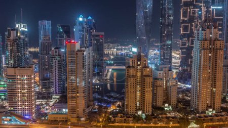 Foto de Rascacielos de Dubai Marina con los edificios residenciales más altos iluminados durante toda la noche timelapse. Canal de agua con botes y luces parpadeantes en ventanas. Vista aérea desde el distrito JLT - Imagen libre de derechos