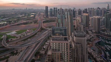 Foto de El puerto deportivo de Dubái y el JLT iluminaron rascacielos a lo largo de Sheikh Zayed Road con tráfico en un gran cruce aéreo noche a día antes del amanecer. Edificios residenciales y de oficinas desde arriba. - Imagen libre de derechos