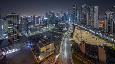 Téléchargez les photos : Panorama montrant Bay Avenue avec des tours modernes éclairées développement résidentiel dans Business Bay ciel nocturne timelapse, Dubaï, Émirats arabes unis. gratte-ciel avec circulation sur une route près d'un grand parking - en image libre de droit