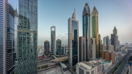 Foto de Panorama aéreo del Distrito Financiero Internacional de Dubai con muchos rascacielos noche a día timelapse transición. Tráfico en una carretera cerca de aparcamiento de varios pisos con piscina en la azotea. Dubai, Emiratos Árabes Unidos. - Imagen libre de derechos