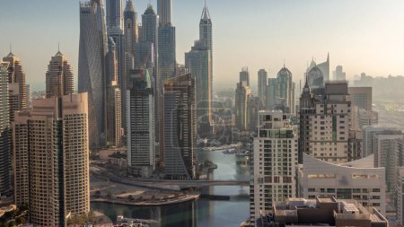 Foto de Vista de varios rascacielos en el bloque recidencial más alto de Dubai Marina durante el amanecer timelapse aéreo con canal artificial. Muchas torres y yates temprano en la mañana con cielo naranja - Imagen libre de derechos