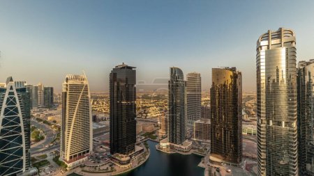 Téléchargez les photos : Bâtiments résidentiels hauts dans le quartier JLT timelapse aérien pendant le coucher du soleil, qui fait partie du centre multi-commodités Dubaï quartier à usage mixte. Gratte-ciel avec de longues ombres et le soleil réfléchi par le verre - en image libre de droit