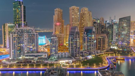 Foto de Dubai Marina con varios barcos cerca de césped verde en el paseo marítimo y rascacielos alrededor de la antena del canal día a la transición nocturna. Torres del distrito JBR en segundo plano - Imagen libre de derechos