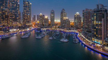 Téléchargez les photos : Baie de yacht de luxe dans la ville transition aérienne de jour à la nuit dans la marina de Dubaï. Des gratte-ciel modernes le long de la promenade riveraine et des bateaux flottant dans le port - en image libre de droit