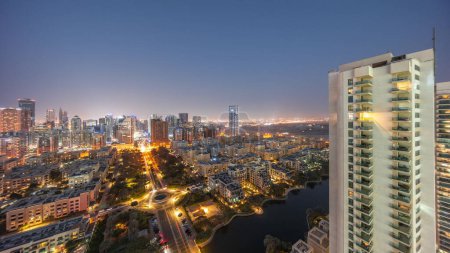 Téléchargez les photos : Des gratte-ciels dans le quartier de Barsha Heights et des immeubles de faible hauteur dans le quartier des Verts transition aérienne de jour à nuit. Skyline de Dubaï avec palmiers et fontaine sur étang - en image libre de droit