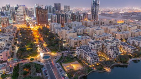 Téléchargez les photos : Bâtiments de faible hauteur dans le quartier des Verts et gratte-ciel dans le quartier de Barsha Heights transition aérienne du jour à la nuit panoramique. Skyline de Dubaï avec palmiers et arbres - en image libre de droit