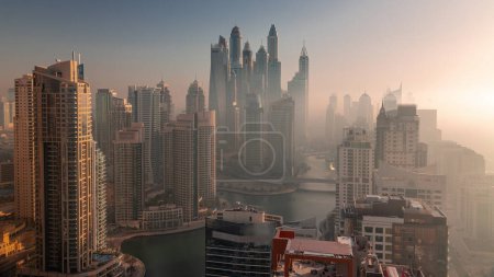 Foto de Vista de varios rascacielos en el bloque recidencial más alto de Dubai Marina durante la salida del sol aérea con canal artificial. Muchas torres y yates temprano niebla mañana - Imagen libre de derechos