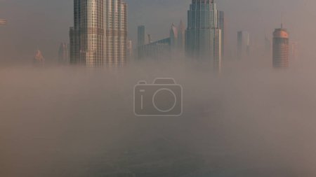 Téléchargez les photos : Vue aérienne de Dubaï tôt le matin pendant le brouillard. Lever de soleil à horizon futuriste de la ville avec des gratte-ciel et des tours d'en haut. Soleil réfléchi par la surface du verre - en image libre de droit