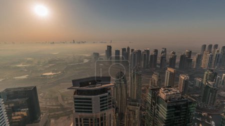 Téléchargez les photos : Panorama de Dubai Marina avec gratte-ciel JLT et parcours de golf au lever du soleil, Dubaï, Émirats arabes unis. Vue aérienne depuis les tours matin brumeux. Skyline de la ville avec ciel orange - en image libre de droit