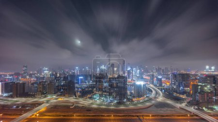 Foto de Skyline panorámico de Dubai con la bahía de negocios y el centro de distrito durante toda la noche. Vista aérea de muchos rascacielos modernos y tráfico ocupado en la carretera al khail. Luna poniéndose y luces apagadas. - Imagen libre de derechos