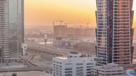 Téléchargez les photos : Site de construction autour du canal d'eau de Dubaï avec de nombreuses grues timelapse aérien. Maisons et villas sont derrière les gratte-ciel de la baie d'affaires. La circulation sur le pont routier. Ciel orange au coucher du soleil - en image libre de droit