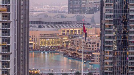 Téléchargez les photos : Vue aérienne de la fontaine de Dubaï au centre-ville avec des palmiers et un drapeau dans le parc à côté du centre commercial et du souk Timelapse de transition du jour à la nuit, Émirats arabes unis. Vu entre deux gratte-ciel d'en haut après le coucher du soleil - en image libre de droit