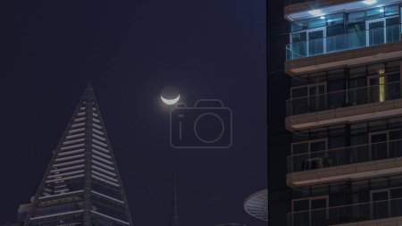 Foto de Crescent Moon está detrás de rascacielos en Dubai timelapse. Majestuosa vista durante la noche en la ciudad. Vista aérea de torres superiores con luz en ventanas. Distrito de Business Bay - Imagen libre de derechos