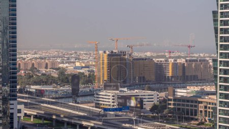 Téléchargez les photos : Site de construction autour du canal d'eau de Dubaï avec de nombreuses grues timelapse aérien. Maisons et villas sont derrière. Trafic sur le pont de l'autoroute le matin avec de longues ombres - en image libre de droit