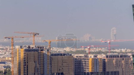 Foto de Sitio de construcción alrededor del canal de agua de Dubái en el paseo marítimo con muchas grúas por la mañana timelapse aérea. Casas y villas están detrás con isla de palmeras. - Imagen libre de derechos