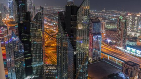 Foto de Edificios de gran altura en Sheikh Zayed Road en Dubai aerial night timelapse, EAU. Rascacielos en el distrito financiero internacional desde arriba. Las casas de paseo de la ciudad y el cruce enorme sobre un fondo - Imagen libre de derechos