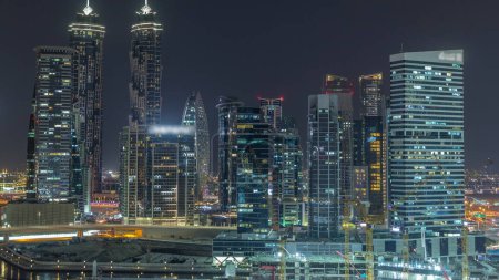 Téléchargez les photos : Paysage urbain avec des gratte-ciel éclairés de Dubai Business Bay et canal d'eau timelapse de nuit aérienne. Skyline moderne de tours résidentielles et de bureaux sur le front de mer. Grues travaillant sur chantier - en image libre de droit