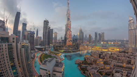 Téléchargez les photos : Gratte-ciel s'élevant au-dessus du centre-ville de Dubaï transition de jour à la nuit, centre commercial et fontaine entourés de bâtiments modernes vue panoramique aérienne avec ciel nuageux au coucher du soleil - en image libre de droit