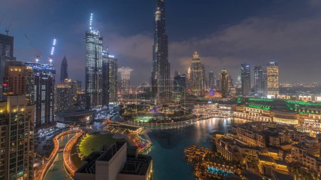 Rascacielos que se elevan por encima del centro de Dubái transición día a noche, centro comercial y fuente rodeada de edificios modernos vista panorámica superior aérea con cielo nublado colorido