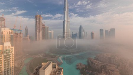 Foto de Vista panorámica aérea de la ciudad de Dubai temprano en la mañana durante la niebla. Salida del sol en el horizonte futurista de la ciudad con rascacielos y torres desde arriba. Sol reflejado desde la superficie de vidrio - Imagen libre de derechos