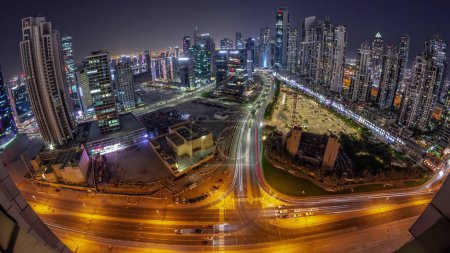 Téléchargez les photos : Skyline panoramique de Bay Avenue avec des tours modernes développement résidentiel dans Business Bay transition aérienne de jour à la nuit, Dubaï, EAU. gratte-ciel avec circulation sur une route près d'un grand parking - en image libre de droit