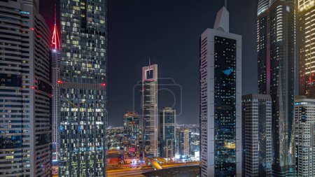 Foto de Panorama que muestra la vista aérea del Distrito Financiero Internacional de Dubái con muchos rascacielos iluminados noche. Tráfico en una carretera cerca de aparcamientos de varios pisos. Dubai, Emiratos Árabes Unidos. - Imagen libre de derechos