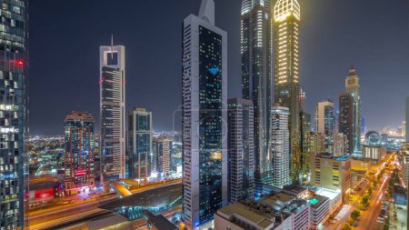 Foto de Aerial view of Dubai International Financial District with many skyscrapers day to night transition after sunset. Traffic on a road near multi storey parking with metro station. Dubai, UAE. - Imagen libre de derechos