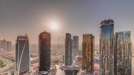 Foto de Salida del sol sobre altos edificios residenciales en la antena del distrito JLT, parte del distrito de uso mixto del centro de productos básicos de Dubai. Rascacielos alrededor del estanque - Imagen libre de derechos