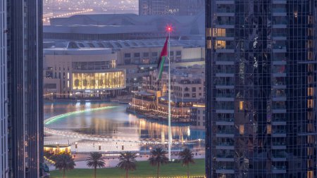 Foto de Vista aérea de Dubai Fuente en el centro con palmeras y bandera en el parque junto al centro comercial y zoco noche a día timelapse transición, Emiratos Árabes Unidos. Visto entre dos rascacielos desde arriba antes del amanecer - Imagen libre de derechos
