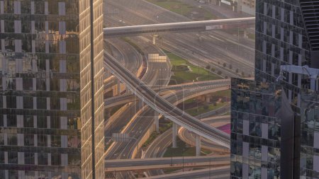 Téléchargez les photos : Vue aérienne entre les gratte-ciel d'en haut à un énorme carrefour routier animé avec ligne de métro dans le centre-ville de Dubaï timelapse pendant le coucher du soleil. Voitures et camions conduisant directement dans les deux sens la route. - en image libre de droit