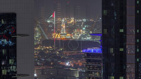 Photo for Aerial view between two skyscrapers to apartment houses and villas in Dubai city near downtown night timelapse. Flag on a flagpole. Illuminated cranes of sea port in a distance. United Arab Emirates - Royalty Free Image