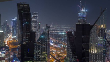 Téléchargez les photos : Vue aérienne d'en haut à un grand carrefour routier animé derrière les gratte-ciel du centre-ville de Dubaï timelapse de nuit. Voitures conduisant directement dans les deux sens. Quartier des affaires avec tours éclairées - en image libre de droit