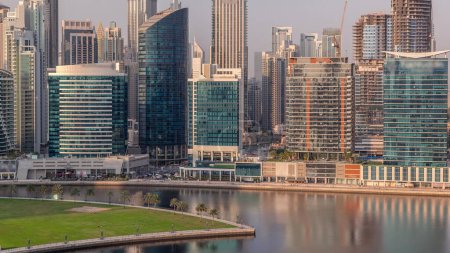 Foto de Vista aérea a Dubai Business Bay y el centro de la ciudad con los diversos rascacielos y torres a lo largo de la costa con palmeras y césped verde en el timelapse canal. Construcción con grúas - Imagen libre de derechos