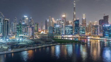 Téléchargez les photos : Vue aérienne vers Dubai Business Bay et le centre-ville avec les différents gratte-ciel et tours le long du front de mer sur le canal timelapse de nuit. Site de construction avec grues sur l'île - en image libre de droit