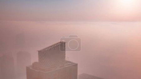 Foto de Salida del sol sobre la rara niebla invernal de la madrugada sobre el horizonte de Dubai Marina y rascacielos iluminados por el orage sun light aerial timelapse. Vista superior desde las nubes superiores. Dubai, Emiratos Árabes Unidos - Imagen libre de derechos