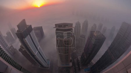 Téléchargez les photos : Rare brouillard hivernal matinal au lever du soleil au-dessus de la ligne d'horizon de Dubai Marina et JLT et des gratte-ciel éclairés par des lampadaires aériens. Vue de dessus depuis les nuages. Dubai, Émirats arabes unis - en image libre de droit