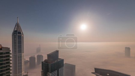 Foto de Salida del sol sobre la rara niebla invernal de la madrugada sobre el horizonte de Dubai Marina y los techos de rascacielos timelapse aéreo. Vista superior desde las nubes superiores. Dubai, Emiratos Árabes Unidos - Imagen libre de derechos