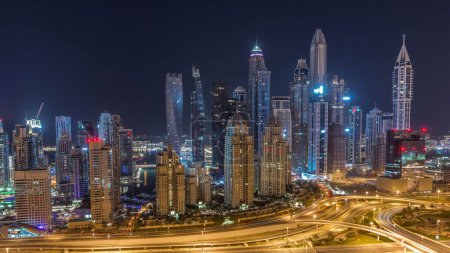 Téléchargez les photos : Les gratte-ciel de Dubai Marina avec les bâtiments résidentiels les plus hauts éclairés pendant toute la nuit timelapse avec des lumières éteintes. Vue aérienne du quartier JLT. Trafic sur une autoroute - en image libre de droit