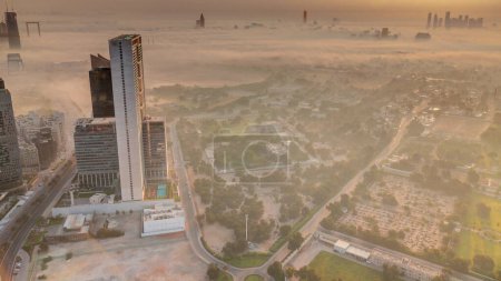 Foto de Villas en el distrito de Zabeel con rascacielos en un timelapse panorámico aéreo de fondo durante el amanecer en Dubai, Emiratos Árabes Unidos. Tráfico en calles con casas y caminos cubiertos por niebla matutina - Imagen libre de derechos
