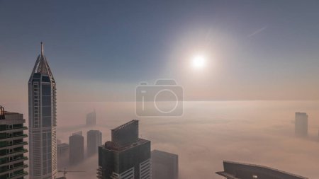 Foto de Salida del sol sobre la rara niebla invernal de la madrugada sobre el horizonte de Dubai Marina y los techos de rascacielos aéreos. Vista superior desde las nubes superiores. Dubai, Emiratos Árabes Unidos - Imagen libre de derechos