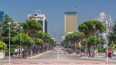Téléchargez les photos : Trafic sur le boulevard Deshmoret e Kombit à Tirana timelapse. Rue principale de la capitale albanaise entourée d'arbres verts et de gratte-ciel. Les voitures se déplacent et s'arrêtent aux intersections avec un feu de circulation - en image libre de droit