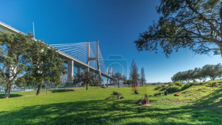 Téléchargez les photos : The Vasco da Gama Bridge timelapse hyperlapse viewed from green lawn with trees. Cable-stayed longest bridge flanked by viaducts and rangeviews that spans Tagus River in Park Nations. Lisbon, Portugal - en image libre de droit