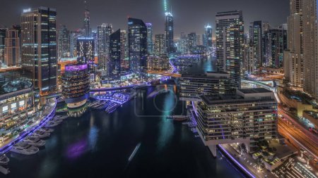 Foto de Panorama que muestra vista aérea al puerto deportivo de Dubai rascacielos iluminados alrededor del canal con yates flotantes noche. Torres en los distritos jlt y jbr. Barcos blancos están estacionados en el club de yates - Imagen libre de derechos