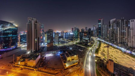 Foto de Canal de agua con torres modernas iluminadas desarrollo residencial en Business Bay panorámica aérea durante toda la noche, Dubai, Emiratos Árabes Unidos. Rascacielos con tráfico en una carretera y luces apagadas - Imagen libre de derechos