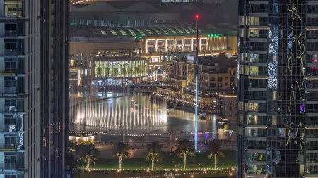 Foto de Vista aérea de la Fuente de Dubai en el centro con palmeras y bandera en el parque junto al centro comercial iluminado y el timelapse de la noche del zoco, Emiratos Árabes Unidos. Visto entre dos rascacielos desde arriba. - Imagen libre de derechos