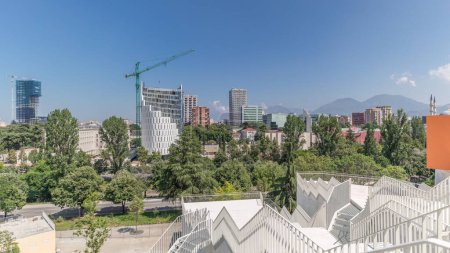 Foto de Panorama que muestra el paisaje urbano de Tirana con sus coloridos edificios de apartamentos y rascacielos timelapse, Tirana, Albania. Vista aérea desde el mirador de la pirámide con árboles verdes y montañas - Imagen libre de derechos