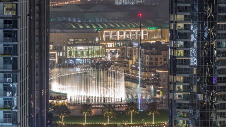 Photo for Aerial view of Dubai Fountain in downtown with palms and flag in park next to illuminated shopping mall and souq night timelapse, UAE. Seen between two skyscrapers from above. - Royalty Free Image