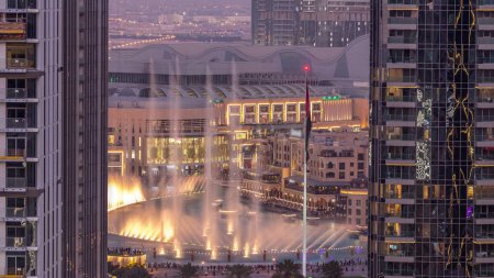 Foto de Vista aérea de Dubai Fuente en el centro con palmeras y bandera en el parque junto al centro comercial y zoco día a noche timelapse transición, Emiratos Árabes Unidos. Visto entre dos rascacielos desde arriba después del atardecer - Imagen libre de derechos